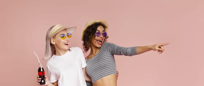 This photo shows two young women smiling and laughing together, looking happy and carefree. The woman on the left is wearing a white t-shirt, sunglasses, and a hat, and holding a bottle of soda