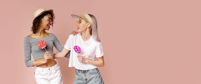 This image shows two women standing in front of a pink backdrop. The women are both wearing sunglasses and hats and holding lollipops. They are laughing and looking at each other.