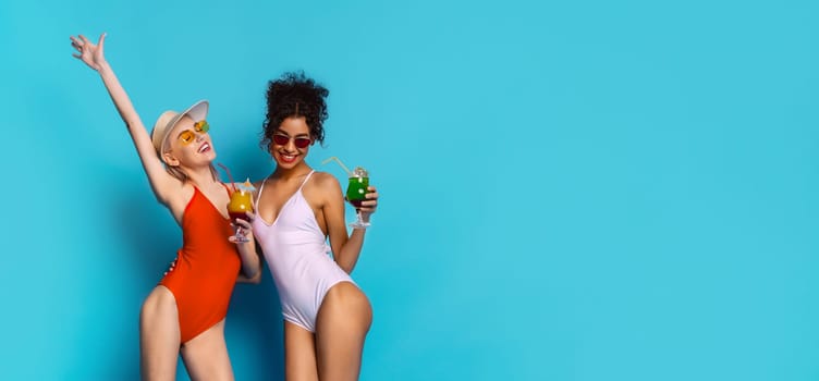 Two women are standing in front of a bright blue background, smiling and holding cocktails. Women are happy and carefree. They look like they are having a great time, copy space