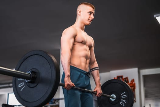 A shirtless young bodybuilder is lifting weights barbell in a gym