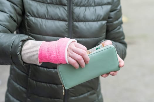 Woman in green coat with pink cast on her arm takes out euro banknotes from an open wallet near a car in a parking lot, High quality photo