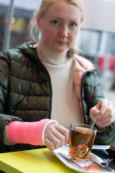 woman with a pink fiberglass cast on her right hand sits in a cafe at a table holding a mug of hot tea in her hand, brewing tea with a teapot,