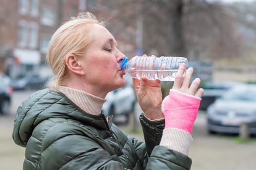 woman in green coat and with a pink fiberglass bandage on her arm drinks water from a bottle in a parking lot in the city, leading an active lifestyle despite restrictions