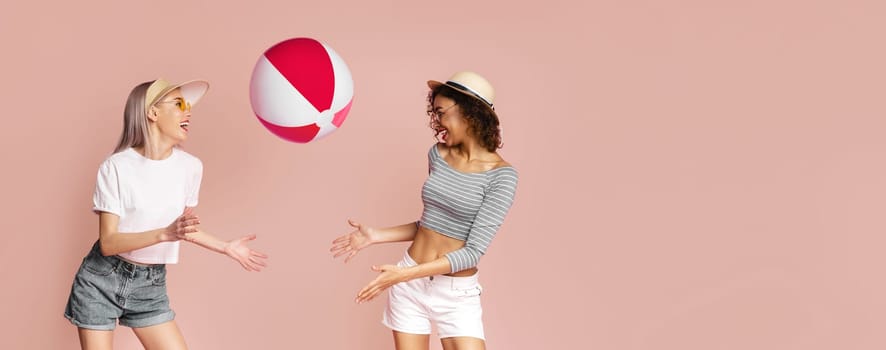 Two young women, one with blonde hair and the other with dark curly hair, are playing with a beach ball in front of a pink background. They are both wearing summery clothing