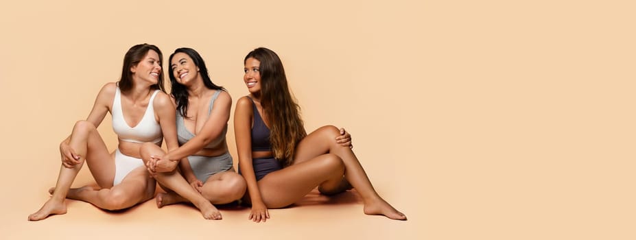 Three glad diverse millennial women in undergarments sit on floor, enjoy body positivity, beauty care, have fun, isolated on light studio background, full length. Friendship, confidence, copy space