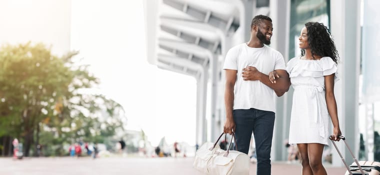 A smiling African American couple walks together outside a modern building on a sunny day. The man has his arm around the womans waist and they are both carrying luggage, copy space