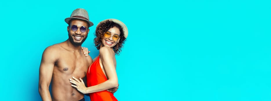 Smiling African American couple wearing sunglasses and hats in front of a bright blue background. The man is shirtless and the woman is wearing a red swimsuit, copy space