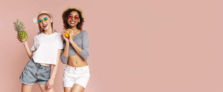 This image shows two young women standing in front of a pink background. They are both smiling and holding fruit, wearing casual summer clothing, including hats and sunglasses, copy space
