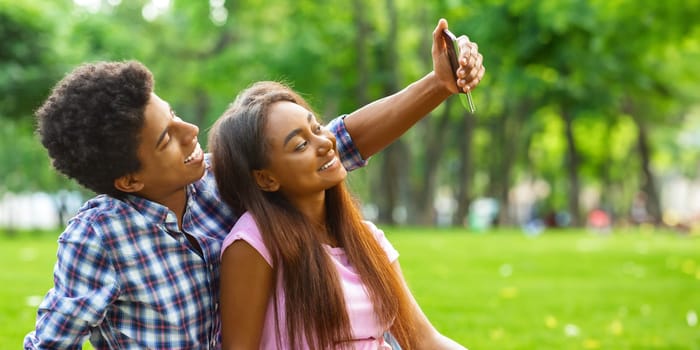 A teen black couple sitting on a grassy field in a park on a sunny day, taking selfie on smartphone. The couple is enjoying the beautiful weather and each others company, copy space
