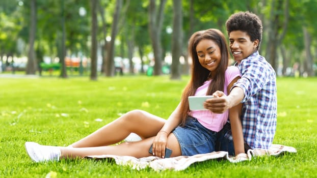A teen black couple sitting on a blanket in a grassy park. Guy is holding a smartphone and taking a selfie with the girl. They are both smiling and looking at the camera