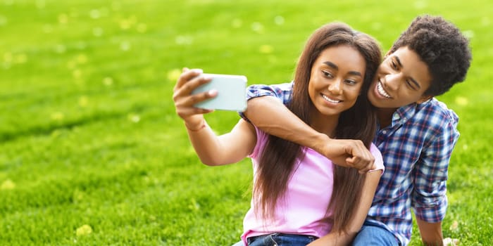A teen black couple is taking a selfie together in a park. They are sitting on the green grass, smiling and looking at the camera, panorama with copy space