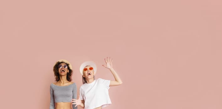 Two young women are laughing at the camera while wearing sunglasses and hats. The background is a solid pink color, copy space