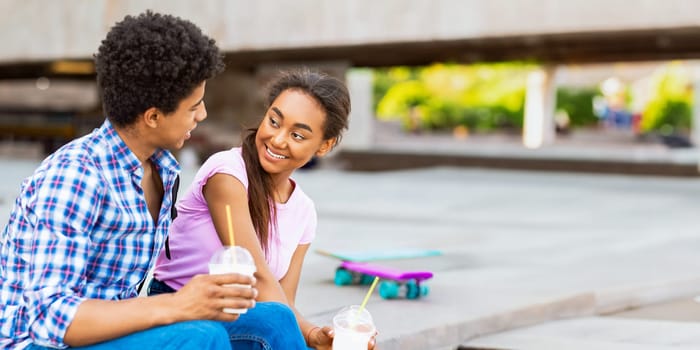 A teen African American couple sits together on a concrete ledge, both holding drinks with straws. They are engaged in conversation, smiling at each other, and enjoying a sunny day, copy space