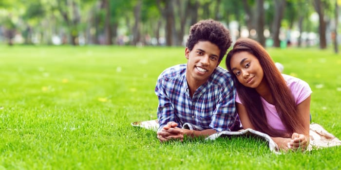 A teen black couple lies on a blanket in a grassy park, surrounded by trees. Guy is wearing a plaid shirt and the girl is wearing a pink t-shirt. They are both smiling and looking at the camera