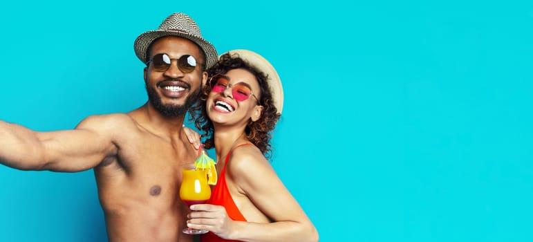 A smiling African American man and woman, wearing sunglasses and hats, stand in front of a blue background. The man is taking a selfie while holding his arm around the woman, copy space