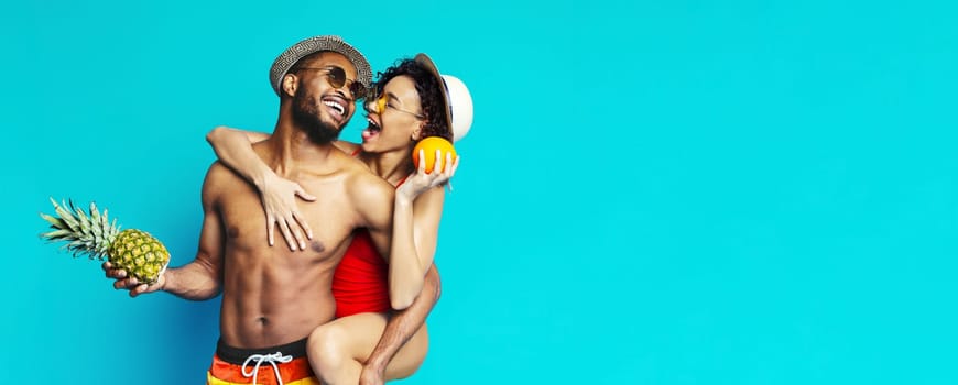 A young couple in swimwear poses in front of a blue background. The man is holding a pineapple and the woman is holding an orange. They are both smiling and looking at each other.