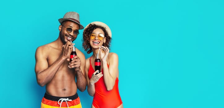 A young black couple dressed in swimwear and sunglasses smiles and enjoys drinks together on a bright blue background.