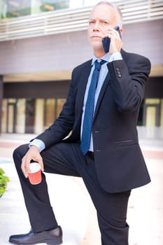 Satisfied with the results, the businessman walks down the street outside the office building, a mature boss holds a phone and coffee in his hands, writes messages and reads news online, using an app.