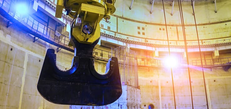 A large, steel claw hangs suspended from a crane inside a vast, concrete structure. The claw appears to be reaching towards a shaft of sunlight that illuminates the room. The structure appears to be under construction and is surrounded by scaffolding and other building materials.