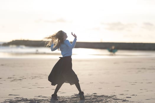 A woman is dancing on the beach in a black skirt. She is wearing a blue shirt and black boots