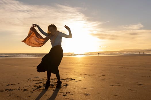 A woman is standing on a rocky hillside, with the sun setting in the background. She is wearing a scarf and she is dancing or performing a ritual. Concept of freedom and connection to nature