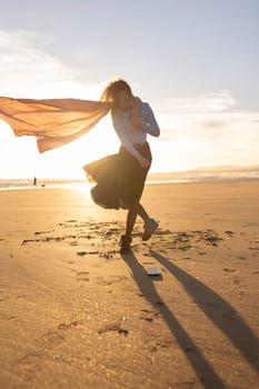 A woman stands on a beach with a scarf blowing in the wind. The scene is serene and peaceful, with the sun setting in the background