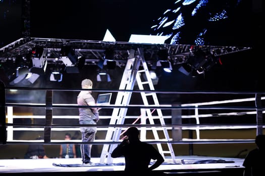 A man is standing in front of a boxing ring with a laptop in his hand. The scene is set in a dark room with a stage and a ring