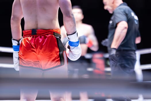 A man in a red boxing shorts is standing in a ring with his hands on his hips. A referee is standing behind him