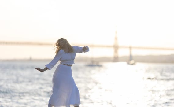 A woman in a white dress is dancing on the beach, with the ocean in the background. She is dancing or doing yoga, and the scene has a peaceful and serene mood
