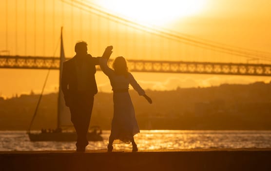 A couple is dancing on a pier by the water. The sun is setting in the background, creating a warm and romantic atmosphere