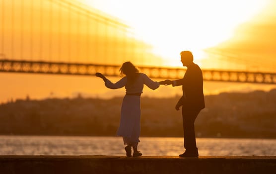 A couple is dancing on a pier by the water. The man is wearing a suit and the woman is wearing a white dress. The sun is setting in the background, creating a warm and romantic atmosphere