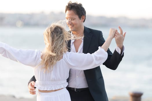 A man and woman are dancing on the beach. The man is wearing a suit and the woman is wearing a white dress. Scene is lighthearted and romantic