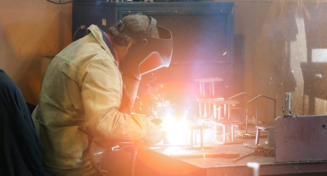 A welder works diligently, wearing a protective mask and gloves, amidst a shower of sparks as they skillfully weld metal pieces. The intense light of the welding torch illuminates the workshop, highlighting the workers concentration.