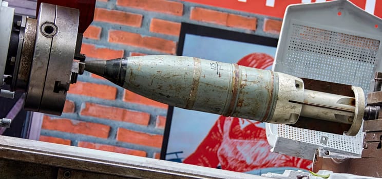 A large, rusted missile is clamped securely within a metal vise. The missile is partially visible, with the front end protruding towards the viewer. The vise is situated in front of a brick wall, with a red and white poster partially visible in the background.