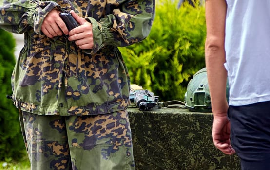 A soldier in full camouflage gear stands with a pistol in his hand, while a civilian walks away in the background. The soldier is focused on his task, while the civilian seems to be unconcerned about the situation. They are surrounded by greenery, suggesting a training exercise or a military event.