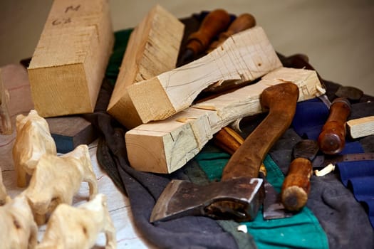 A small group of carved wooden animals stands on a workbench, emerging from the chaos of a woodworker's workshop. A large axe and various tools are scattered across the surface, hinting at the intricate process that brought the animal figures to life. A piece of unfinished wood is still being worked on, suggesting that more creations are yet to be born.