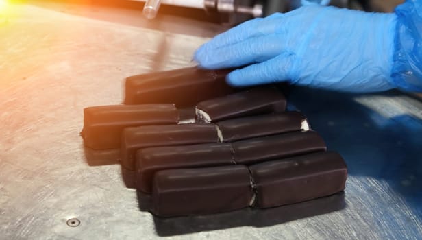A gloved hand inspects a batch of freshly made chocolate bars on a stainless steel surface at a confectionery production facility. Each bar is aligned uniformly, awaiting the next phase of packaging or additional quality checks to ensure consistency and excellence.