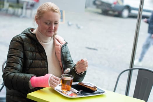 woman with a pink plaster cast on her right arm sits in a cafe and drinks tea with eclair, active life despite restrictions