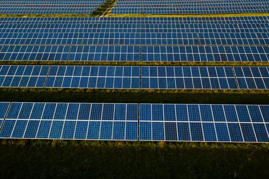 Aerial view of large field filled with rectangular solar panels, resembling patchwork of dark green rectangles against light green grass and asphalt paths