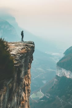 Person standing on a high cliff edge with a breathtaking view of a river and mountains during sunset