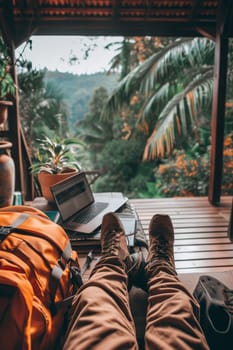 Close-up of a traveler's legs with a laptop on a wooden balcony, overlooking a lush jungle setting