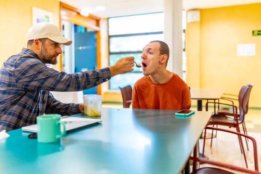 Man helping to eat to a disabled man in a cowokring cafeteria