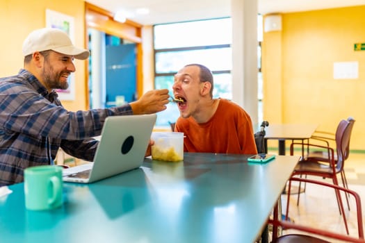 Caucasian male colleague feeding a man with disability in a coworker cafeteria