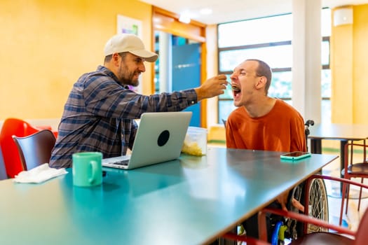 Man feeding a coworker with disability in the public cafeteria of a modern coworking