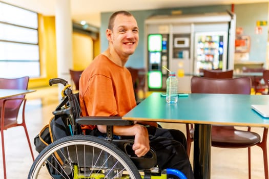 Three quarter length photo of a smiling caucasian man with disability sitting drinking water in the university cafeteria