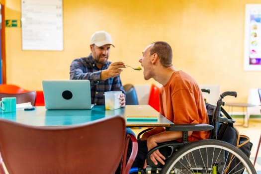Colleague feeding a man with cerebral palsy in a coworking cafeteria in a modern space