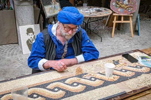 dinan,france,28-04-2024:a man sits at a table and makes art by putting pieces of stone together like mosaics to create beautiful images like paintings