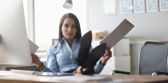 Portrait of cute lady in company office. Overworked businesslady holding biz document and looking at camera with calmness. Businesswoman sitting on chair and putting feet on table