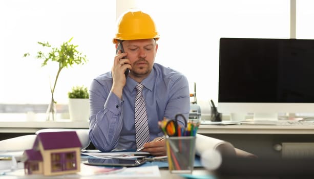 Caucasian Engineer Working on House Blueprint. Portrait of Bearded Architect Sitting at Workplace. Man Wearing Shirt and Yellow Hard Hat make Phone Call. Layout Plan of Rooms Lay on Desk