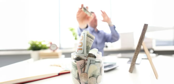 Rich man throwing US banknotes in big jar full of money at workplace closeup